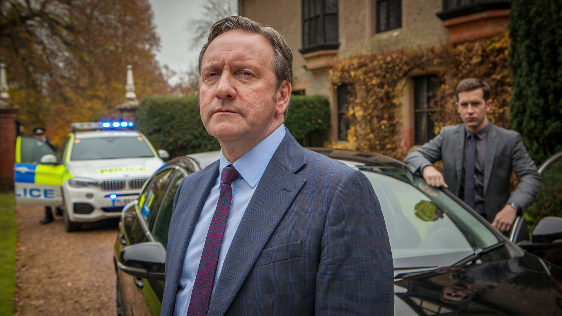 "Inspector Barnaby - Die verfluchte 9": DCI John Barnaby (Neil Dudgeon) und sein Assistent Jamie Winter (Nick Hendrix) stehen neben ihrem Wagen. Im Hintergrund parkt ein Streifenwagen.