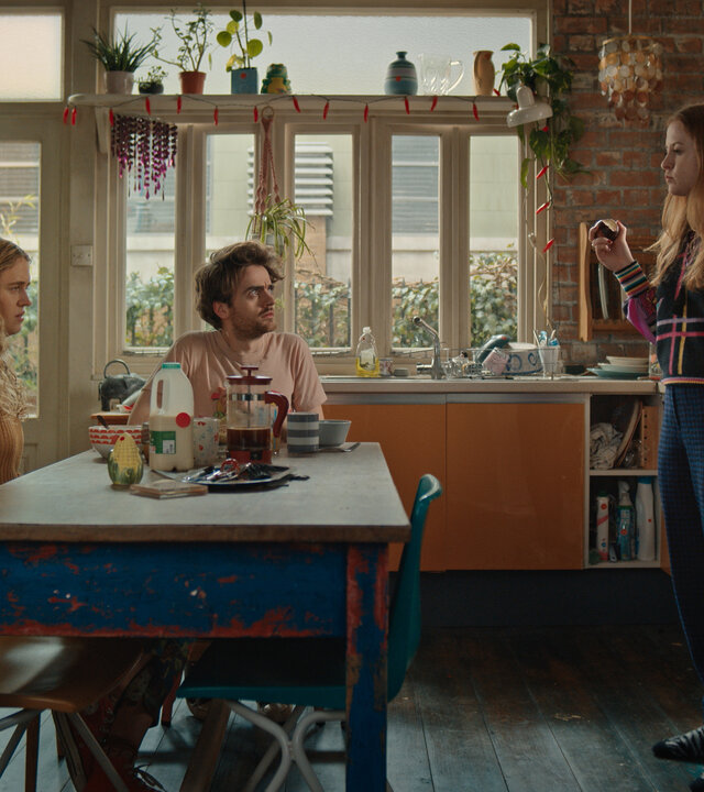 "Extraordinary - Unsere Freunde sind die wahren Superkräfte": Carrie (Sofia Oxenham), Jizzlord (Luke Rollason) und Jen (Máiréad Tyers) sind gemeinsam in der Küche. Jen steht, Carrie und Jizzlord sitzen am gedeckten Tisch. Jen beißt demonstrativ in ihre Zwiebel.
