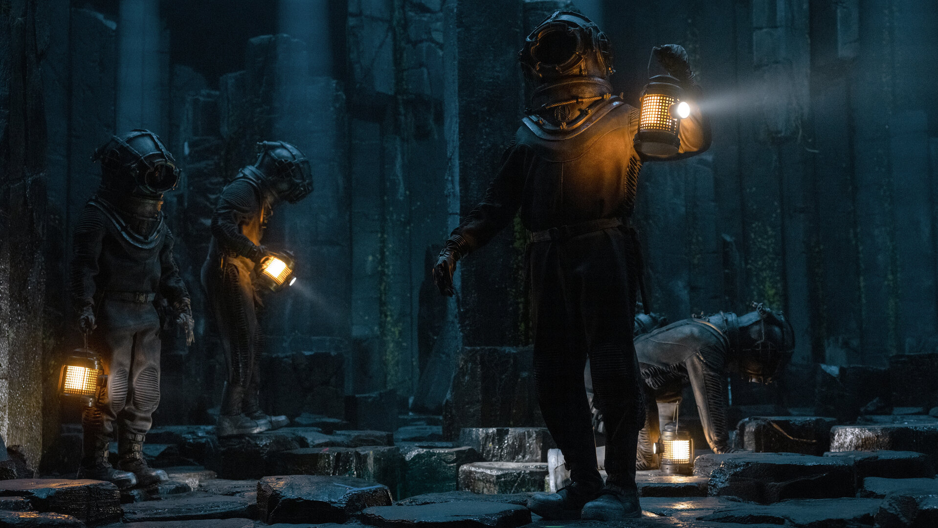 "Nautilus - Das große Blau": Humility (Georgia Flood), Nemo (Shazad Latif), Benoit (Thierry Frémont) und Cuff (Benedict Hardie) sind in einer dunklen Umgebung unter Wasser unterwegs. Alle vier tragen Tiefseetaucheranzüge und Helme. Cuff kniet auf dem Boden und hat seine Leuchte abgestellt. Die anderen laufen herum und halten ihre Leuchten in den Händen.