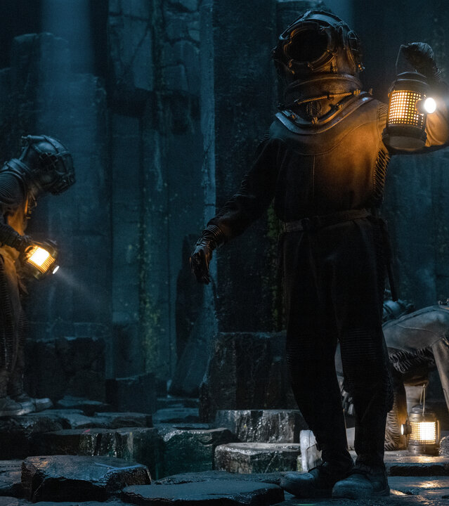 "Nautilus - Das große Blau": Humility (Georgia Flood), Nemo (Shazad Latif), Benoit (Thierry Frémont) und Cuff (Benedict Hardie) sind in einer dunklen Umgebung unter Wasser unterwegs. Alle vier tragen Tiefseetaucheranzüge und Helme. Cuff kniet auf dem Boden und hat seine Leuchte abgestellt. Die anderen laufen herum und halten ihre Leuchten in den Händen.