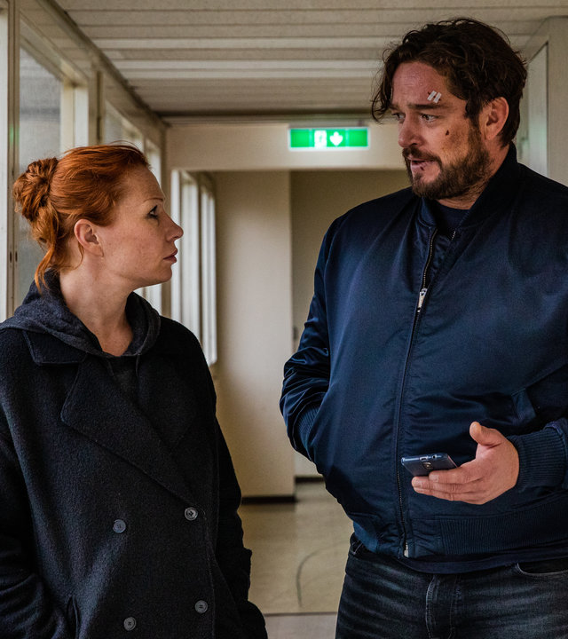 "Dengler - Kreuzberg Blues": Olga Illiescu (Birgit Minichmayr) und Georg Dengler (Ronald Zehrfeld) stehen einander zugewandt im Fenstergang eines Hochhauses. Olga schaut Dengler aufmerksam an, während er ihr etwas erzählt und sein Handy vor sich in der Hand hält. Im Gesicht hat er Verletzungen.
