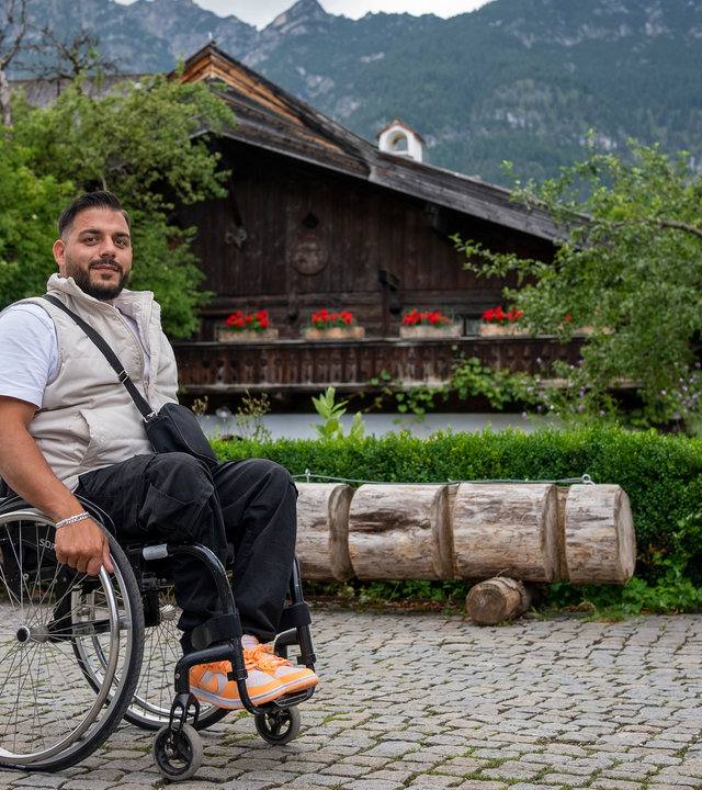 "einfach Mensch - Komm ich zeig Dir meine Heimat - Oberbayern": Ein junger Mann sitzt in seinem Rollstuhl im Freien auf einem gepflasterten Platz. Im Hintergrund ist ein Brunnen aus Holz, ein Holzhaus und Berge.