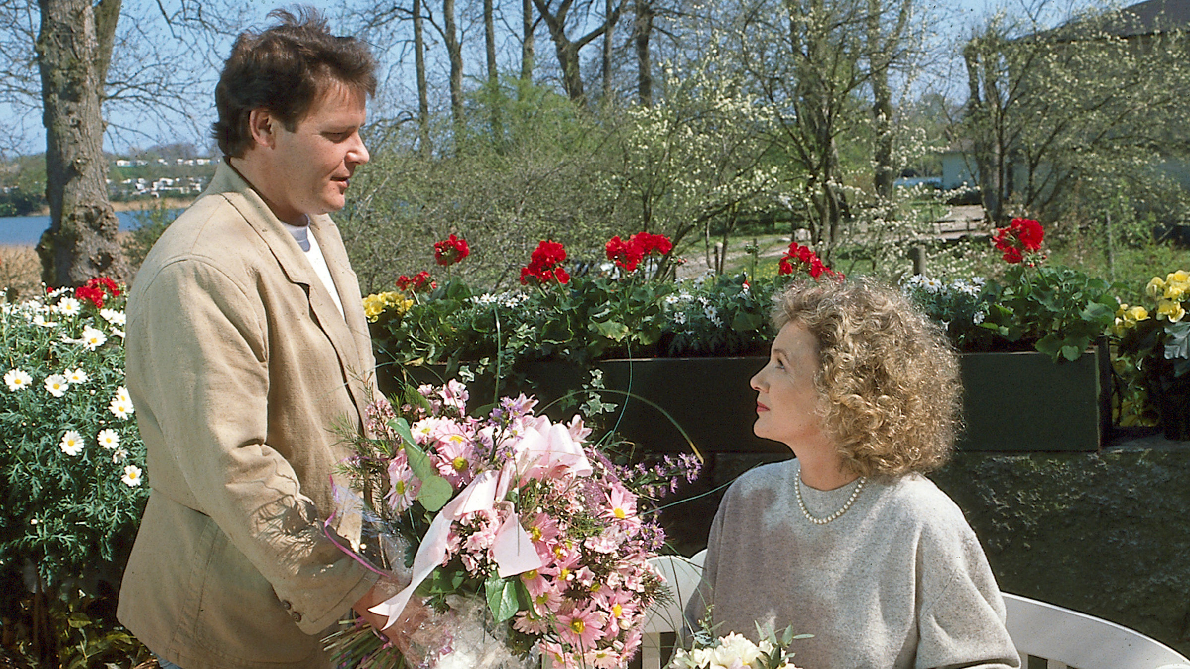 "Der Landarzt - Der Held von Deekelsen": Dr. Teschner (Walter Plathe) und Annemarie Mattiesen (Gila von Weitershausen) draußen auf einer Terrasse, im Hintergrund rote Geranien und weiße Magariten. Dr. Teschner steht lächelnd vor der sitzenden Annemarie mit einem großen rosafarbenen Blumenstrauß. Sie sieht lächelnd zu ihm hoch.