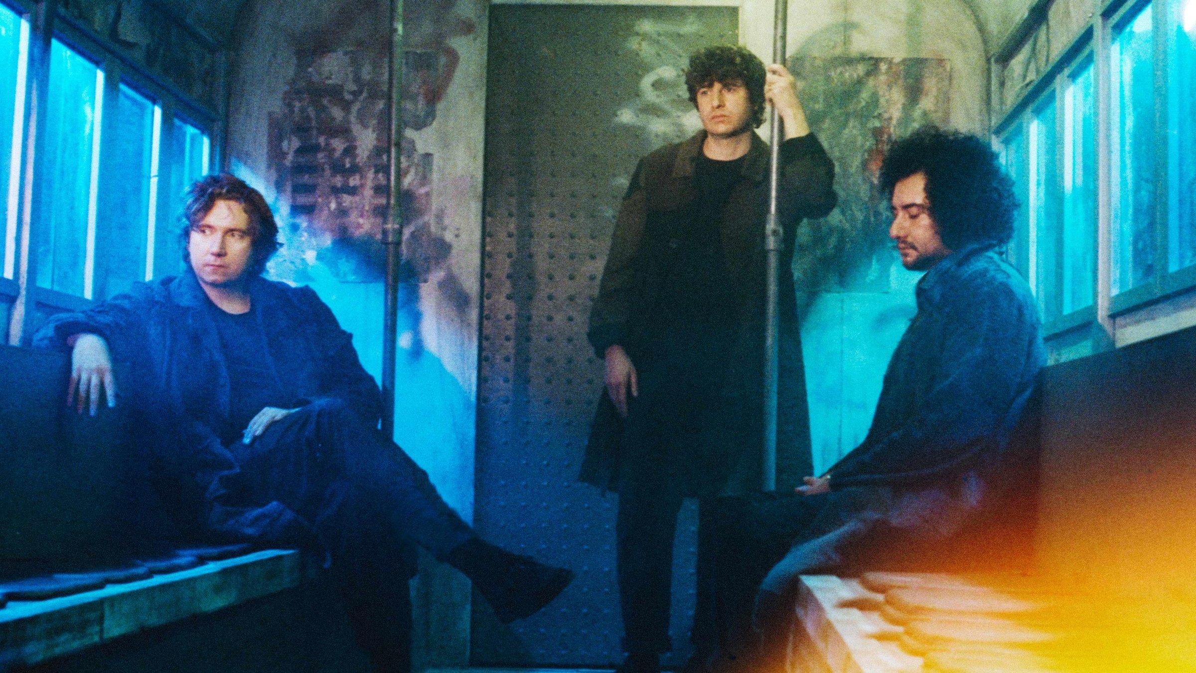 "Pop Around @ Bauhaus: The Kooks": Drei Männer in schwarzer Kleidung sind in einem uralten U-Bahn-Waggon. Einer sitzt links und lehnt sich lässig auf die Rückenlehne der Sitzbank, einer sitzt rechts und guckt geradeaus, der dritte steht leicht angelehnt an einer Haltestange und schaut direkt in die Kamera.