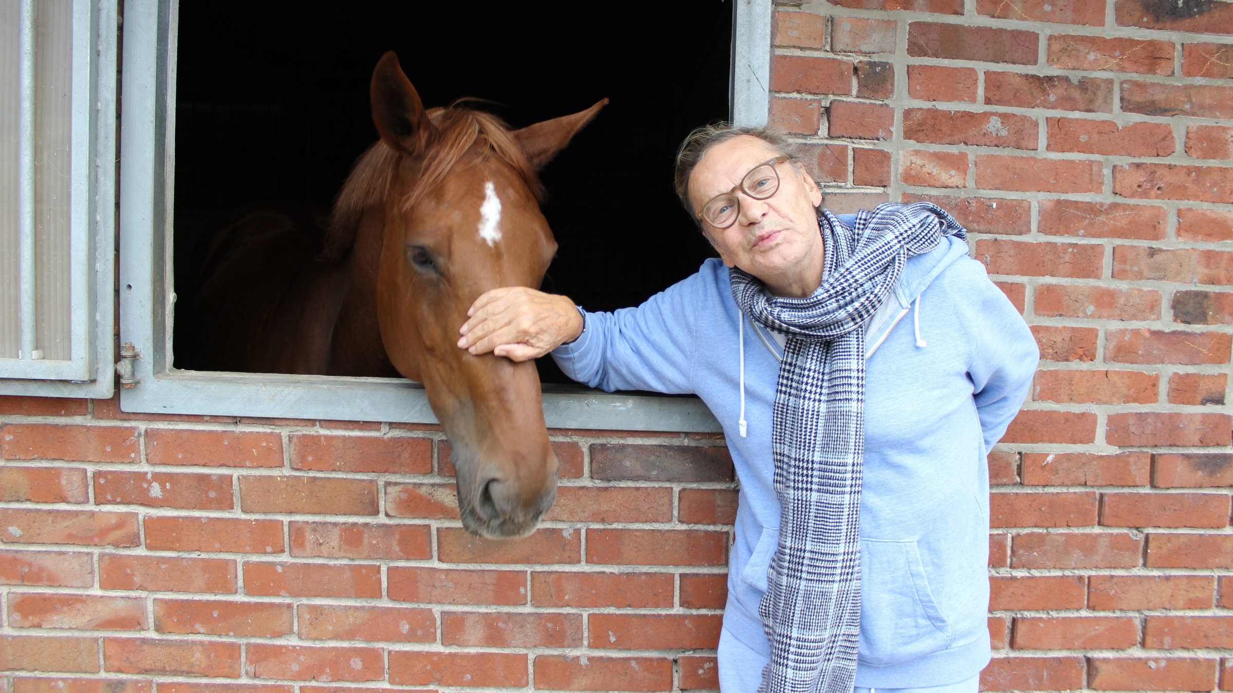"Helmut Berger, meine Mutter und ich": Helmut Berger steht an der Außenwand eines aus Steinen gemauerten Stalls. Aus dem großen Fenster des Stalls schaut ein braunes Pferd heraus. Berger hat seine Hand liebevoll auf den Nasenrücken des Pferdes gelegt und lächelt in die Kamera.