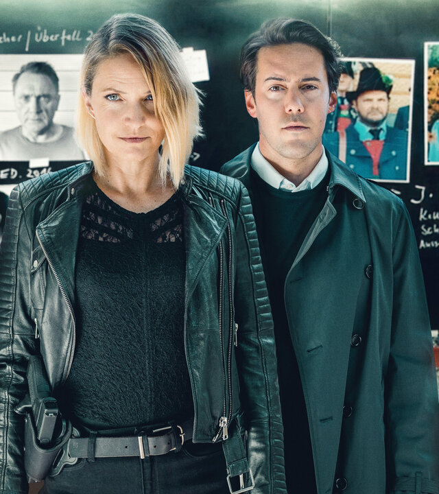 "Der Tote in der Schlucht": Lisa Kuen (Patricia Aulitzky) und Alexander Yüsüf-Demir (Dominik Raneburger) stehen vor einer Tafel mit Fotos und handschriftlichen Notizen. Beide schauen ernst in die Kamera. Die Tafel enthält Bilder von mehreren Personen sowie Diagramme und Textfragmente.
