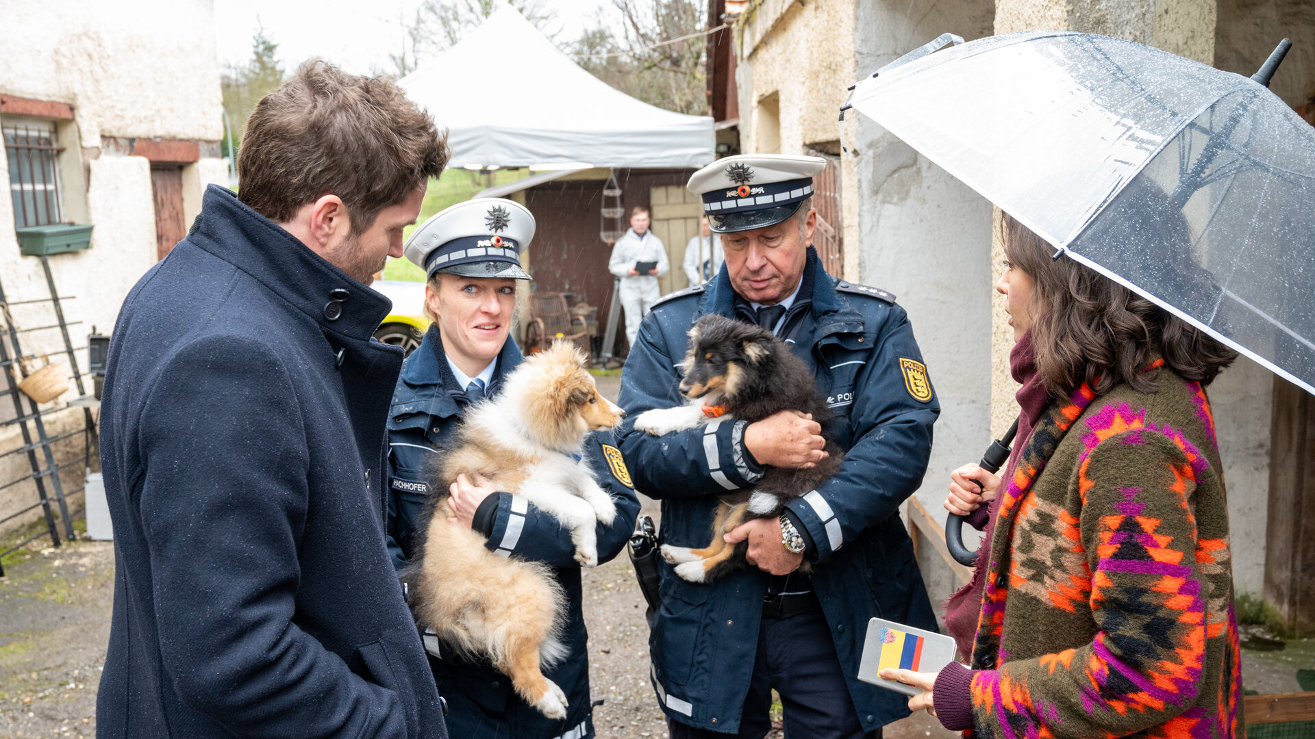 "SOKO Stuttgart - Hundeelend": Die Kommissare Lea Gomez (Jasmin Gassmann) und Jo Stoll (Peter Ketnath) stehen in der Einfahrt eines Hofguts zwei uniformierten Beamten (Komparsen) gegenüber. Jeder der Beamten hält einen Hundewelpen auf dem Arm. Lea hat in der einen Hand einen Regenschirm, in der anderen ein Notizbuch.