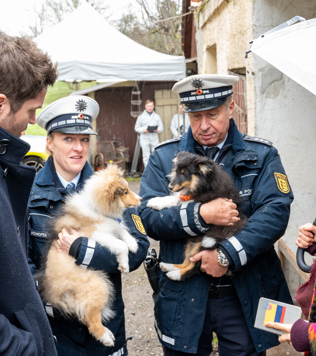 "SOKO Stuttgart - Hundeelend": Die Kommissare Lea Gomez (Jasmin Gassmann) und Jo Stoll (Peter Ketnath) stehen in der Einfahrt eines Hofguts zwei uniformierten Beamten (Komparsen) gegenüber. Jeder der Beamten hält einen Hundewelpen auf dem Arm. Lea hat in der einen Hand einen Regenschirm, in der anderen ein Notizbuch.
