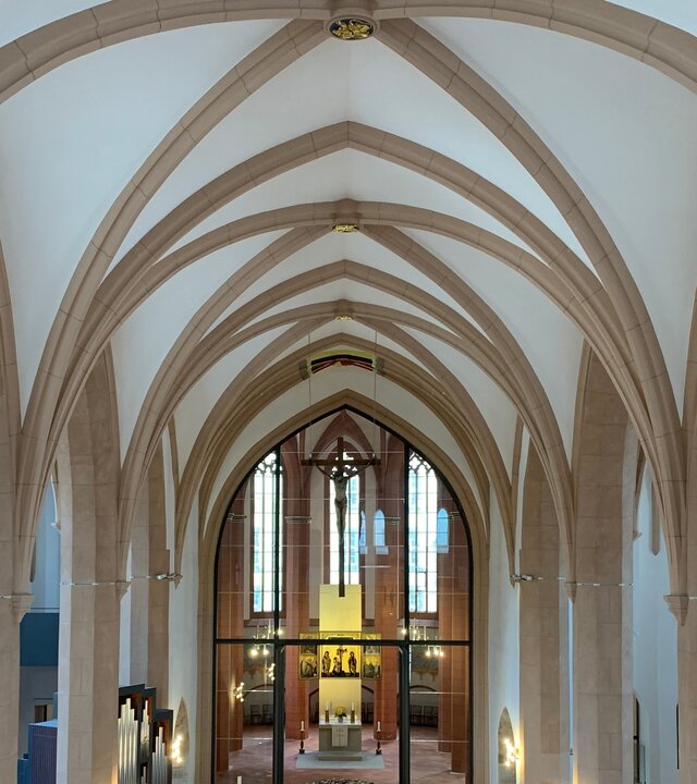 "Evangelischer Gottesdienst - Das Ungesehene sehen": Von oben sind die hellen Säulen und Bögen der St. Jakobi-Kirche in Chemnitz fotografiert. In der hinteren Bildmitte befindet  sich der Altarraum, der in ein warmes Licht getaucht ist. Über ihm hängt ein filigranes Kreuz von der Decke.