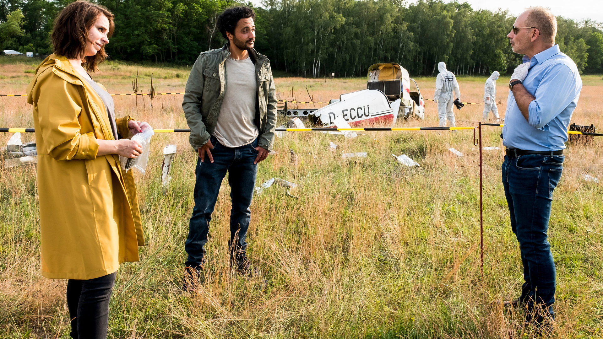 "SOKO Potsdam - Flug in den Tod": Luna Kunath (Caroline Erikson), David Grünbaum (Omar El-Saeidi) und Bernhard Henschel (Michael Lott) stehen auf einem Provinzflugplatz, der als Tatort abgesperrt ist. Bernhard Henschel trägt Handschuhe sowie eine Sonnenbrille und hält eine Hand an seine Brust. David Grünbaum schaut gelassen zu ihm. Im gelben Regenmantel hält Luna Kunath ein Beweisstück in einer Tüte. Die Spurensicherung im Hintergrund sichert das abgestürzte Sportflugzeug ab.