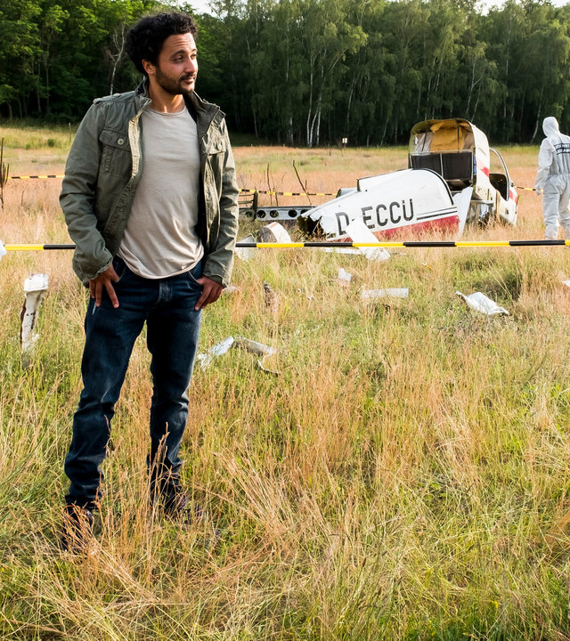 "SOKO Potsdam - Flug in den Tod": Luna Kunath (Caroline Erikson), David Grünbaum (Omar El-Saeidi) und Bernhard Henschel (Michael Lott) stehen auf einem Provinzflugplatz, der als Tatort abgesperrt ist. Bernhard Henschel trägt Handschuhe sowie eine Sonnenbrille und hält eine Hand an seine Brust. David Grünbaum schaut gelassen zu ihm. Im gelben Regenmantel hält Luna Kunath ein Beweisstück in einer Tüte. Die Spurensicherung im Hintergrund sichert das abgestürzte Sportflugzeug ab.