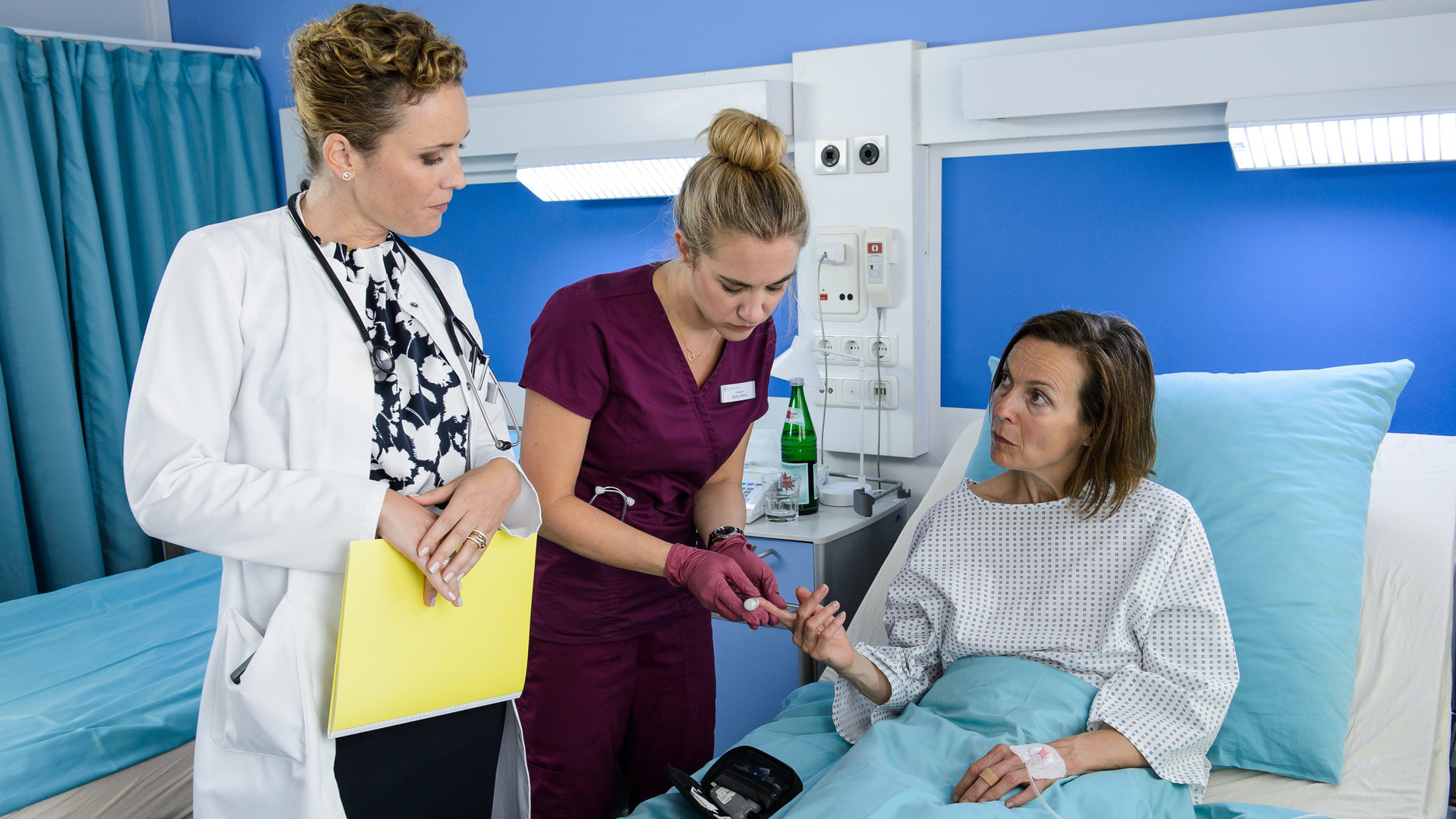 "Bettys Diagnose - Bewährungsprobe": Dr. Helena von Arnstett (Claudia Hiersche) beobachtet Schwester Betty (Annina Hellenthal) genau, während sie der im Krankenbett liegenden Patientin Hilde Rosenbauer (Caroline Schreiber) am Finger Blut abnimmt.