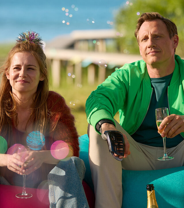 "Dr. Nice: Alte Narben": Charlie (Josefine  Preuß)  und Neiss (Patrick Kalupa) sitzen neben einander auf roten und türkisen Sitzsäcken im Garten, halten ein Glas Sekt in der Hand und lächeln. Um sie herum fliegen Seifenblasen.