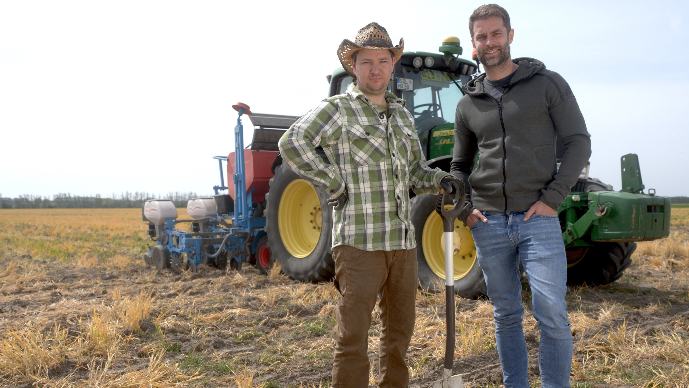 "ZDFzeit: Trocknet Deutschland aus? Gefahren, Fakten, Lösungen": Vor einem Traktor stehen Landwirt Michel Allmrodt und Kai Kupferschmidt auf einem Acker. Michel Allmrodt trägt ein kariertes Hemd, einen Hut und stützt sich auf einen Spaten, Kai Kupferschmidt hat beide Hände in den Hosentaschen.