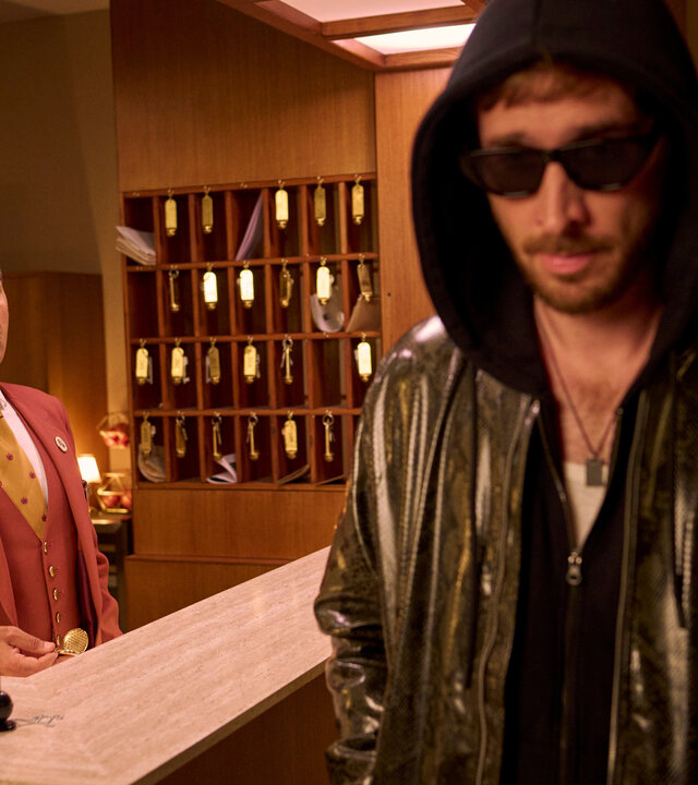 "Nighties - Die Nacht mit dem VIP": Ein Ryan Gosling Double (Tim Vanwersch) läuft in Lederjacke, Kapuzenpullover und Sonnenbrille an einer Hotelrezeption vorbei, hinter der Hotelrezeption steht Charles (Adnan Maral). Hinter ihm steht ein Schlüsselregal, auf dem Tresen liegt eine Klingel und ein Halter mit Stift.