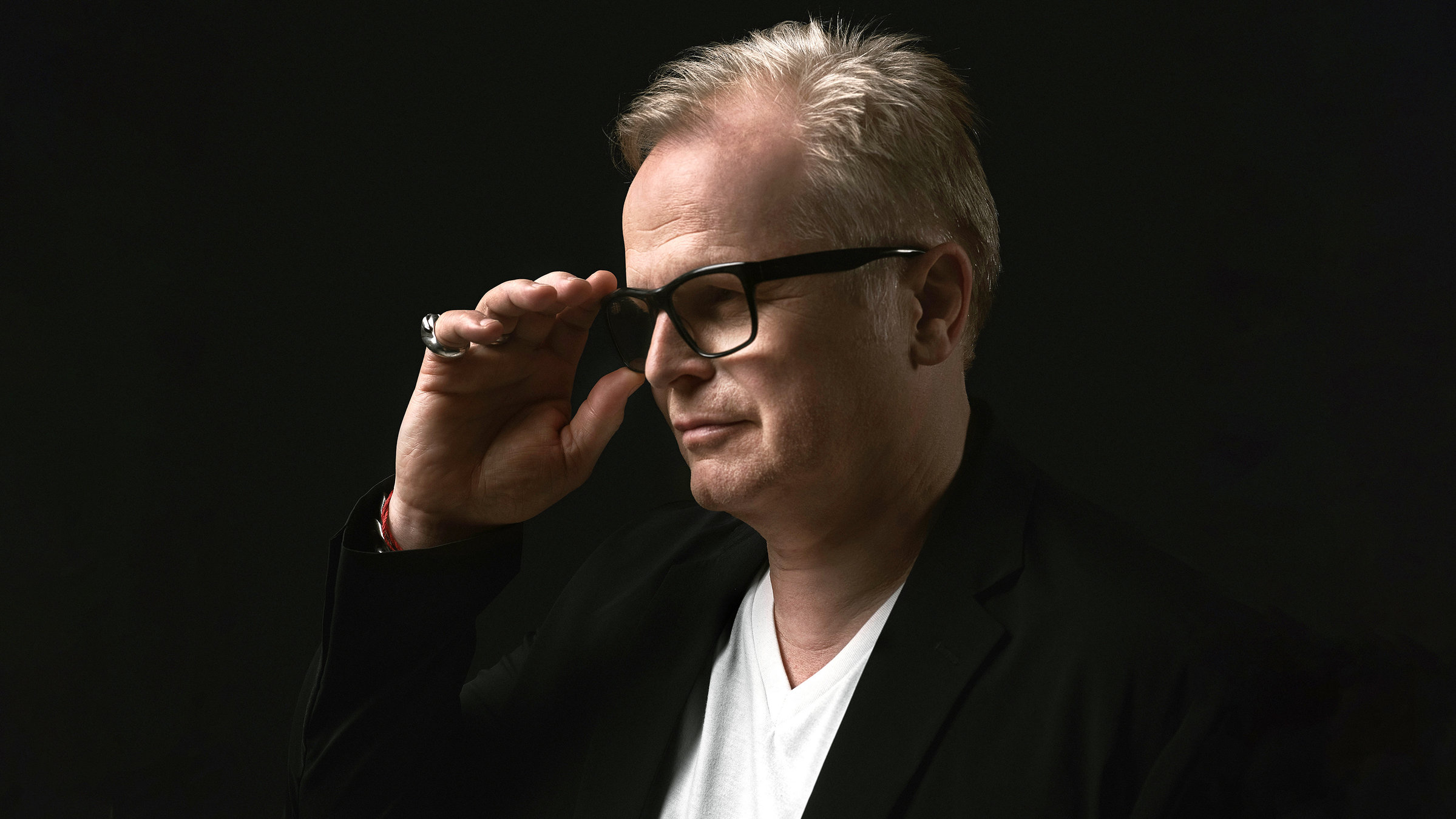 "Herbert Grönemeyer: Mensch - Live": Herbert Grönemeyer steht vor einem schwarzen Hintergrund. Er blickt seitlich an der Kamera vorbei. Mit der rechten Hand fasst er an seine schwarzgeränderte Brille. Bekleidet ist er mit einem schwarzen Jacket und einem weißen T-Shirt.