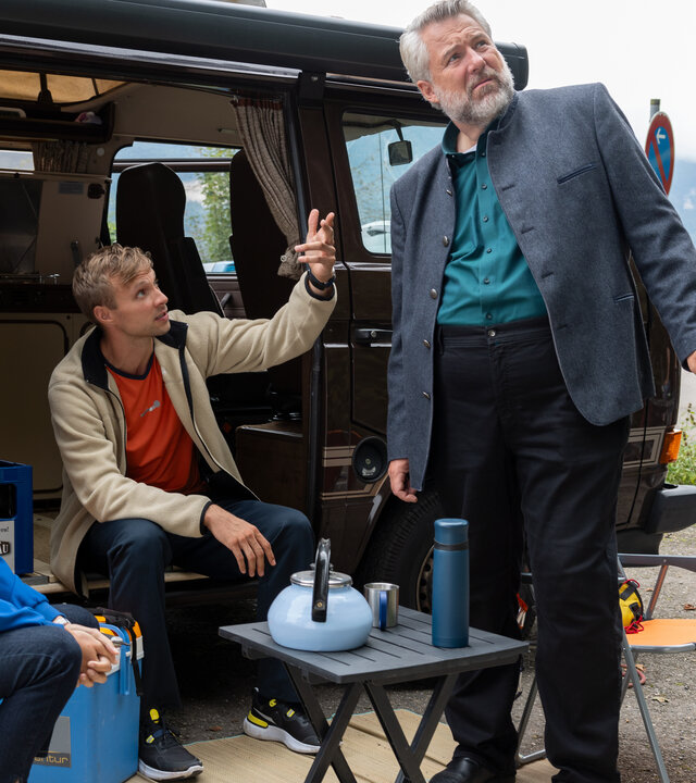 "Die Rosenheim-Cops - Am seidenen Faden": Hauptkommissarin Beck (Michaela Weingartner) sitzt auf einem orangefarbenen Campingstuhl und sieht Fritz Heidenreich (Benedikt Schulz) an, der in der offenen Schiebetür seines dunklen Campingbusses sitzt und nach oben schaut, während er gestikuliert. Neben ihm steht Hauptkommissar Stadler (Dieter Fischer), der in dieselbe Richtung schaut. Vor ihnen steht ein dunkler Klapptisch, auf dem ein hellblauer Teekessel steht. Im Hintergrund parkt ein Polizeifahrzeug an einem asphaltierten Weg.