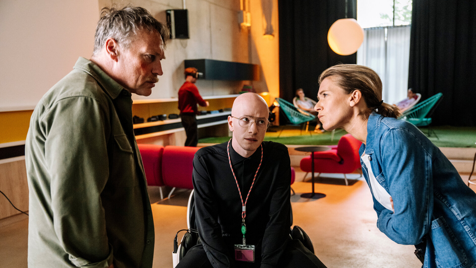 "SOKO Köln - Ewige Jugend": Vanessa Haas (Kerstin Landsmann) und Matti Wagner (Pierre Besson) stehen sich in einem hellen Innenraum gegenüber. Vanessa hat die Arme vor der Brust verschränkt, lehnt sich nach vorne und sieht ihren Kollegen streng an. Matti erwidert ernst ihren Blick. Zwischen ihnen sitzt Theo Sommerfeld (Markus J. Bachmann) und sieht nachdenklich zu Vanessa. Im Hintergrund sitzen zwei Personen (Komparsen) auf türkisen Lounge-Sesseln, während eine weitere Person (Komparse) vor einem roten Sofa steht.