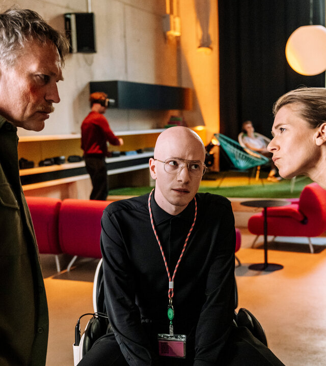 "SOKO Köln - Ewige Jugend": Vanessa Haas (Kerstin Landsmann) und Matti Wagner (Pierre Besson) stehen sich in einem hellen Innenraum gegenüber. Vanessa hat die Arme vor der Brust verschränkt, lehnt sich nach vorne und sieht ihren Kollegen streng an. Matti erwidert ernst ihren Blick. Zwischen ihnen sitzt Theo Sommerfeld (Markus J. Bachmann) und sieht nachdenklich zu Vanessa. Im Hintergrund sitzen zwei Personen (Komparsen) auf türkisen Lounge-Sesseln, während eine weitere Person (Komparse) vor einem roten Sofa steht.