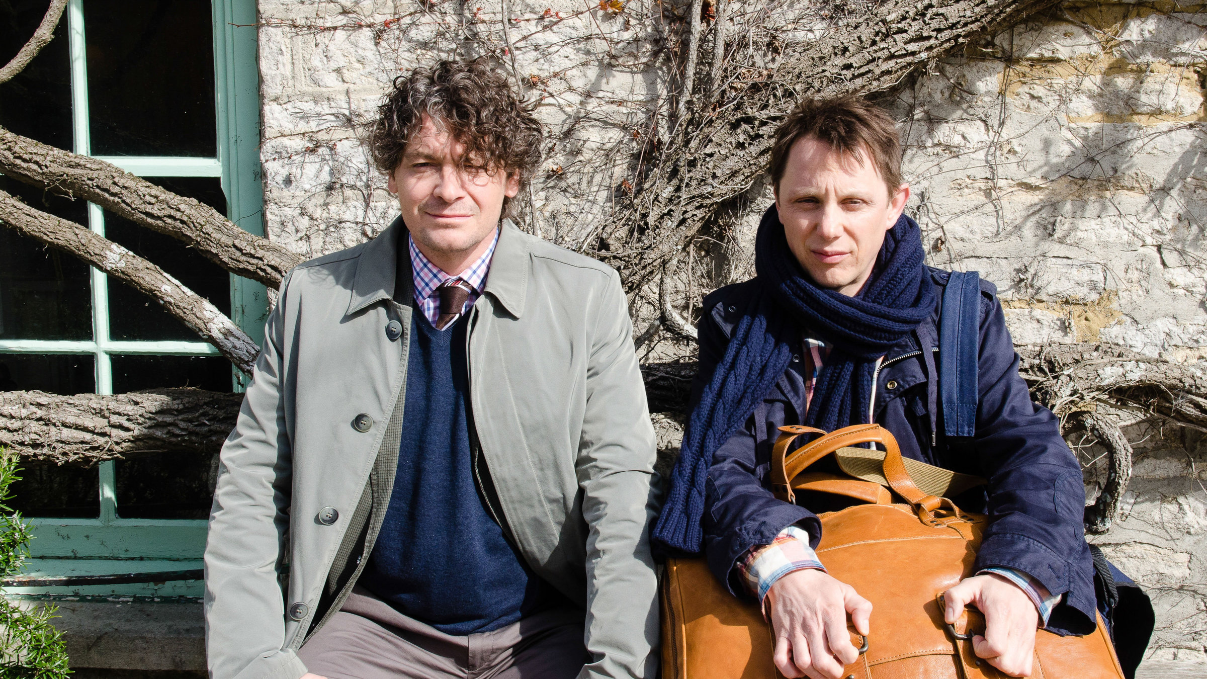 "Inspektor Jury - Der Tote im Pub": Fritz Karl (Inspektor Jury), Arndt Schwering-Sohnrey (Alfred Wiggins) sitzen vor einem Haus nebeneinander auf einer Bank und schauen ernst aus dem Bild.