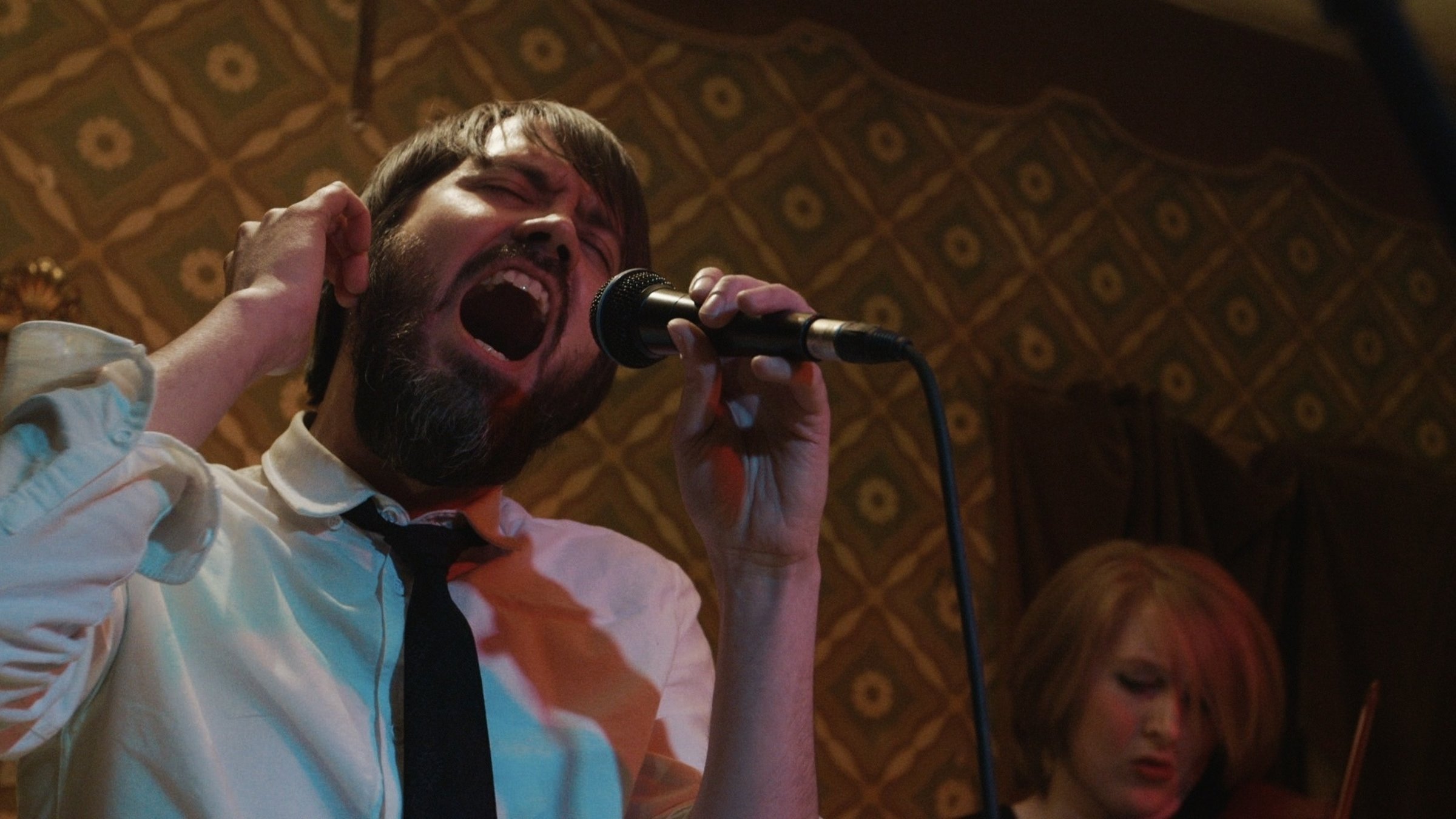 "Wie ein Fremder - Eine deutsche Popmusik-Geschichte - Home":  Junger, bärtiger Mann halbnah, der mit geschlossenen Augen in ein Mikrophon singt, im Hintergrund der Kopf einer jungen Frau.