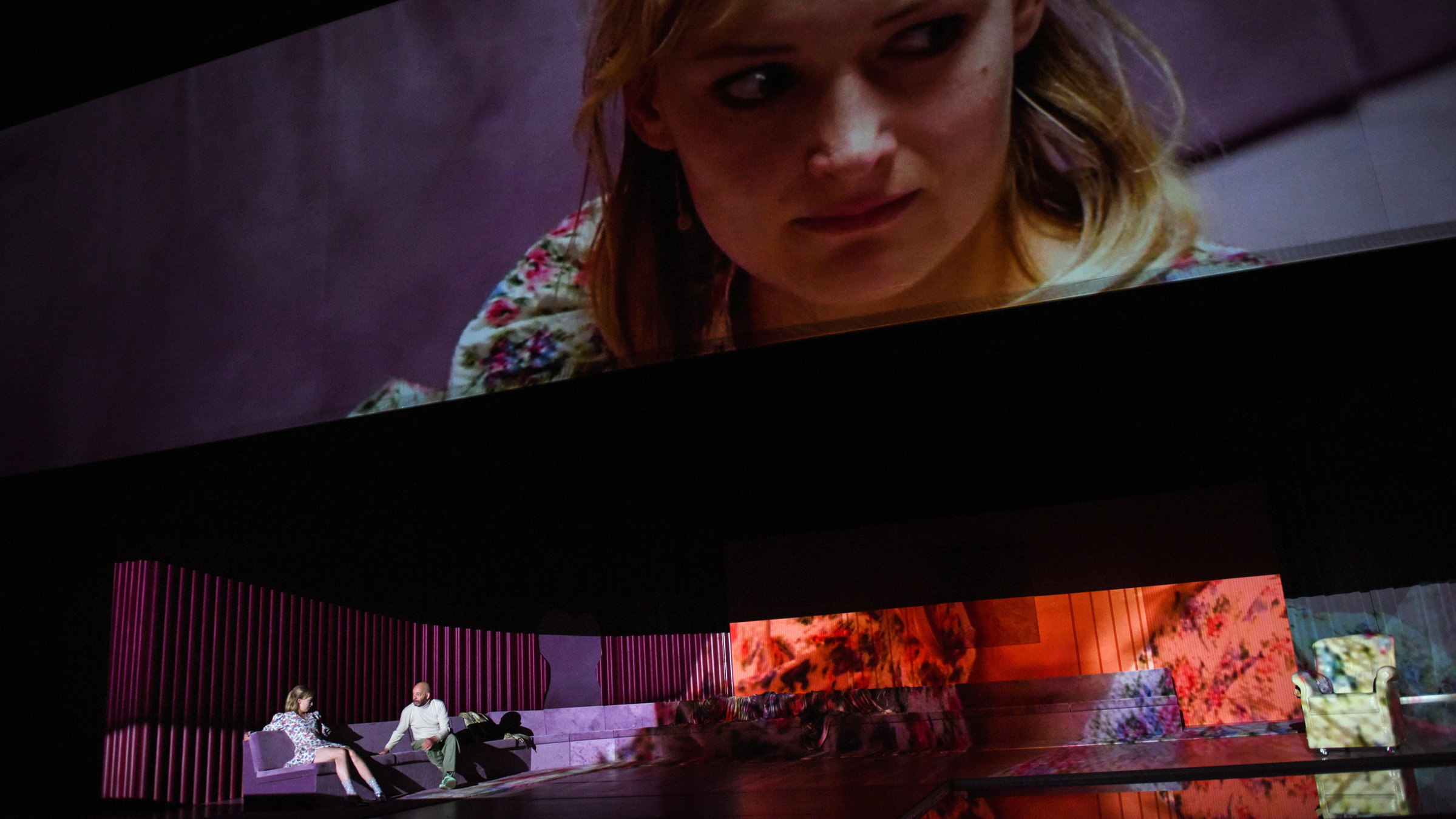 "Starke Stücke: Die Jungfrau von Orleans": Auf einem riesigen, lilafarbenen Ecksofa, das sich fast um die ganze Bühne zieht, sitzen eine junge, blonde Frau in einem weißen Kleid mit Blümchenmuster und ein glatzköpfiger Mann in hellbrauner Hose und weißem Rollkragenpullover. Rechts auf der Bühne steht ein gelber Sessel. Über der Bühne befindet sich eine Cinemascope-Leinwand, auf der das Gesicht der jungen Frau von Nahem zu sehen ist.