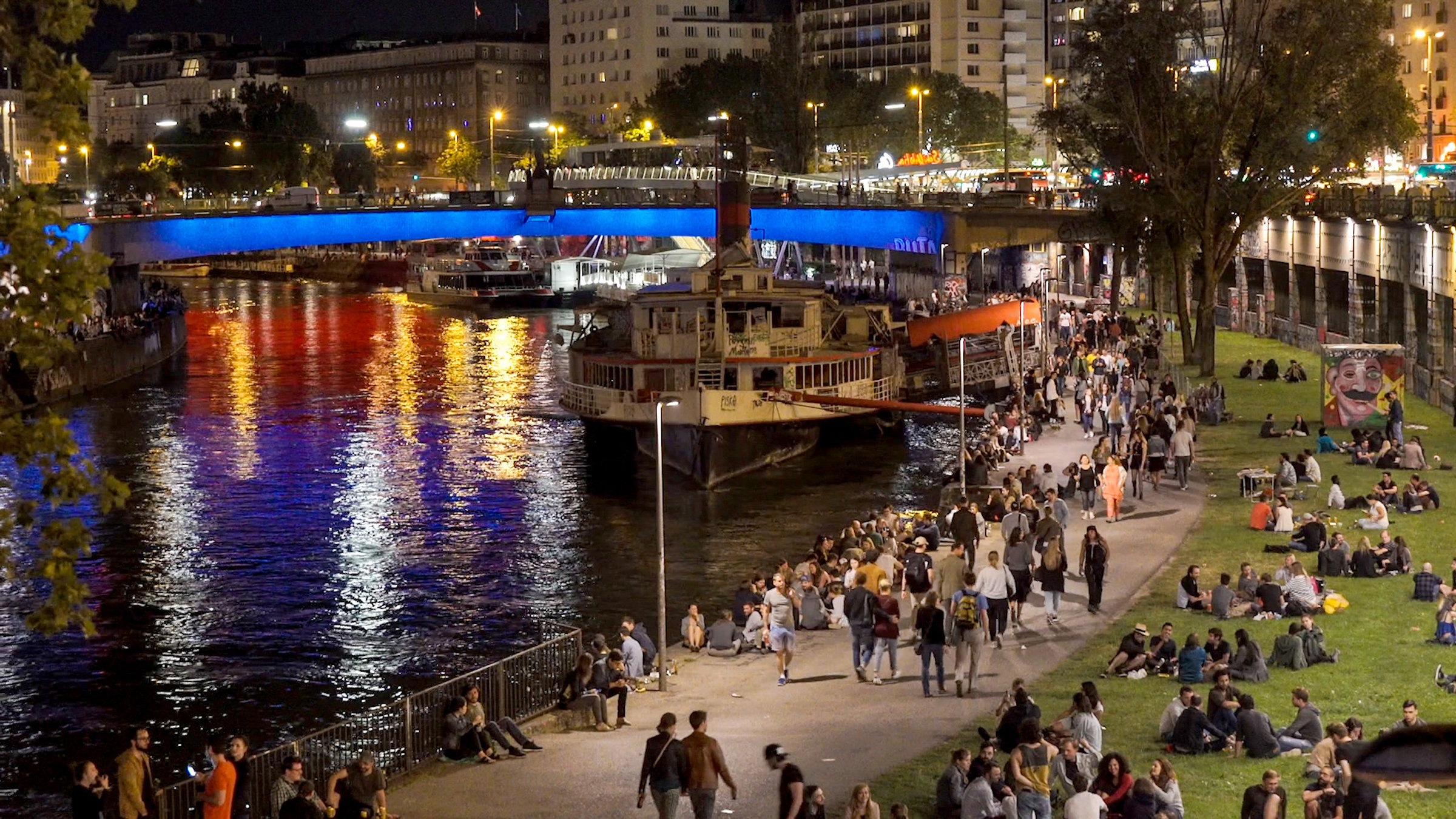 "Wiener Wässer - Der Donaukanal": Menschen flanieren am Ufer am Abend, im Hintergrund eine blau beleuchtete Brücke