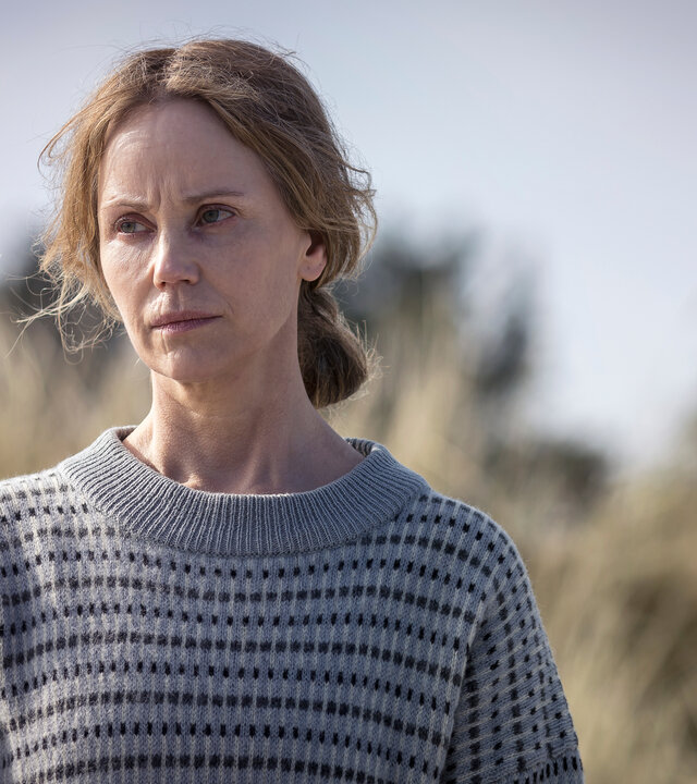 "Iris - Die Wahrheit – Flucht": Iris (Sofia Helin) steht vor einer Uferlandschaft und blickt nachdenklich nach vorne. Sie trägt einen gemusterten, grauen Pullover.