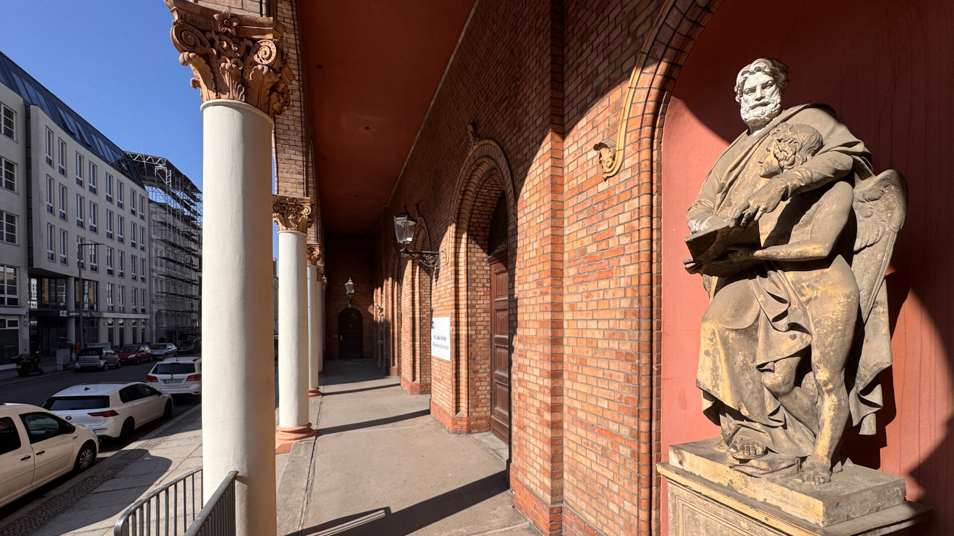 "Evangelischer Gottesdienst - Angekommen": Der Arkadengang, der aus rotem Stein erbauten St. Lukas Kirche inmitten der Berliner Friedrichvorstadt, wird von weißen Säulen gestützt. Vorne im Gang befindet sich eine Statue des Evangelisten Matthäus mit einem Engel. An der Seite parken Autos vor Stadthäusern.