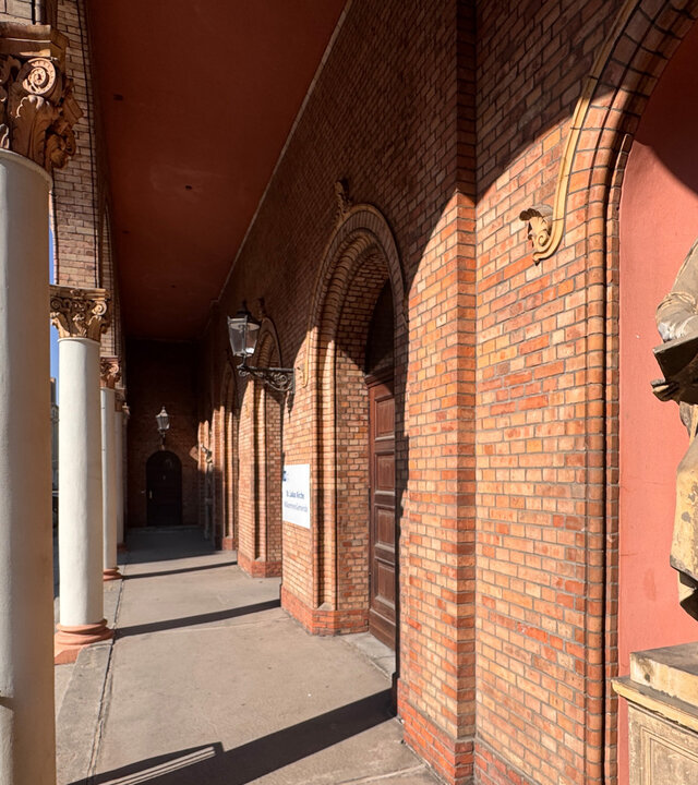 "Evangelischer Gottesdienst - Angekommen": Der Arkadengang, der aus rotem Stein erbauten St. Lukas Kirche inmitten der Berliner Friedrichvorstadt, wird von weißen Säulen gestützt. Vorne im Gang befindet sich eine Statue des Evangelisten Matthäus mit einem Engel. An der Seite parken Autos vor Stadthäusern.