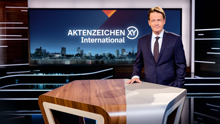 "Aktenzeichen XY... International": Rudi Cerne steht im Studio und lächelt in die Kamera.