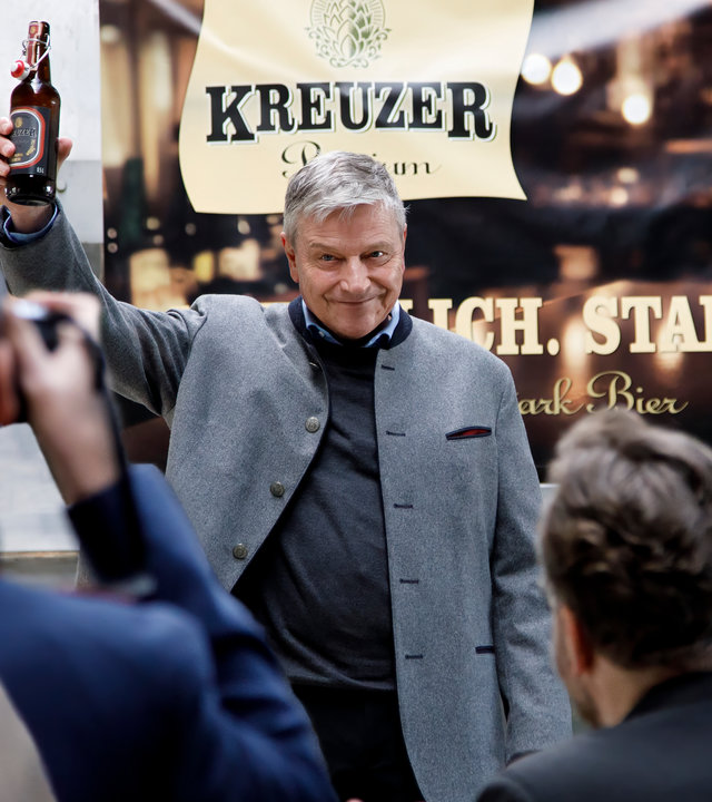 "SOKO Potsdam - Prost": In der Brauerei Kreuzer steht Georg Kreuzer (Christian Maria Goebel) vor einer Versammlung und hält eine Flasche seiner neuen Biersorte "Kreuzer/Stark" in die Luft.