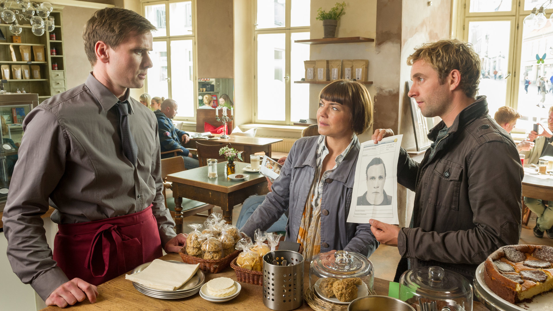 "SOKO Wismar - Durchzug": Katrin Börensen (Claudia Schmutzler) und Nils Theede (Jonas Laux) zeigen dem Kellner (Marco Braun) ein Phantombild. Kellner ist durch eine rote Schürze, die er trägt, erkennbar. Im Hintergrund an den Tischen sind  zu die Besucher des Cafés zu sehen, die von Komparsen gespielt werden.