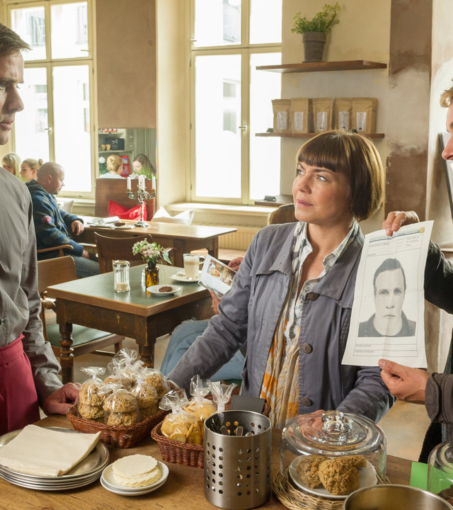 "SOKO Wismar - Durchzug": Katrin Börensen (Claudia Schmutzler) und Nils Theede (Jonas Laux) zeigen dem Kellner (Marco Braun) ein Phantombild. Kellner ist durch eine rote Schürze, die er trägt, erkennbar. Im Hintergrund an den Tischen sind  zu die Besucher des Cafés zu sehen, die von Komparsen gespielt werden.