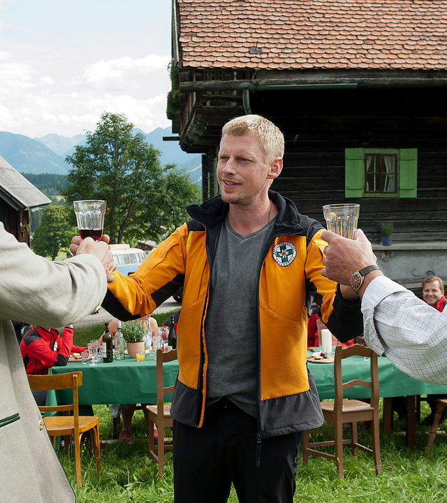 "Die Bergretter: Der Tote im Berg (2)": Andreas (Martin Gruber) steht auf dem Rasen vor einem Haus. Er hat beide Arme ausgestreckt und hält Franz (Heinz Marecek) und Peter (Michael König) ein gefülltes Glas hin. Diese stehen ihm gegenüber und blicken sich mit ernster Miene an. Im Hintergrund sitzt die Crew des Rettungsteams an einem gedeckten Tisch.