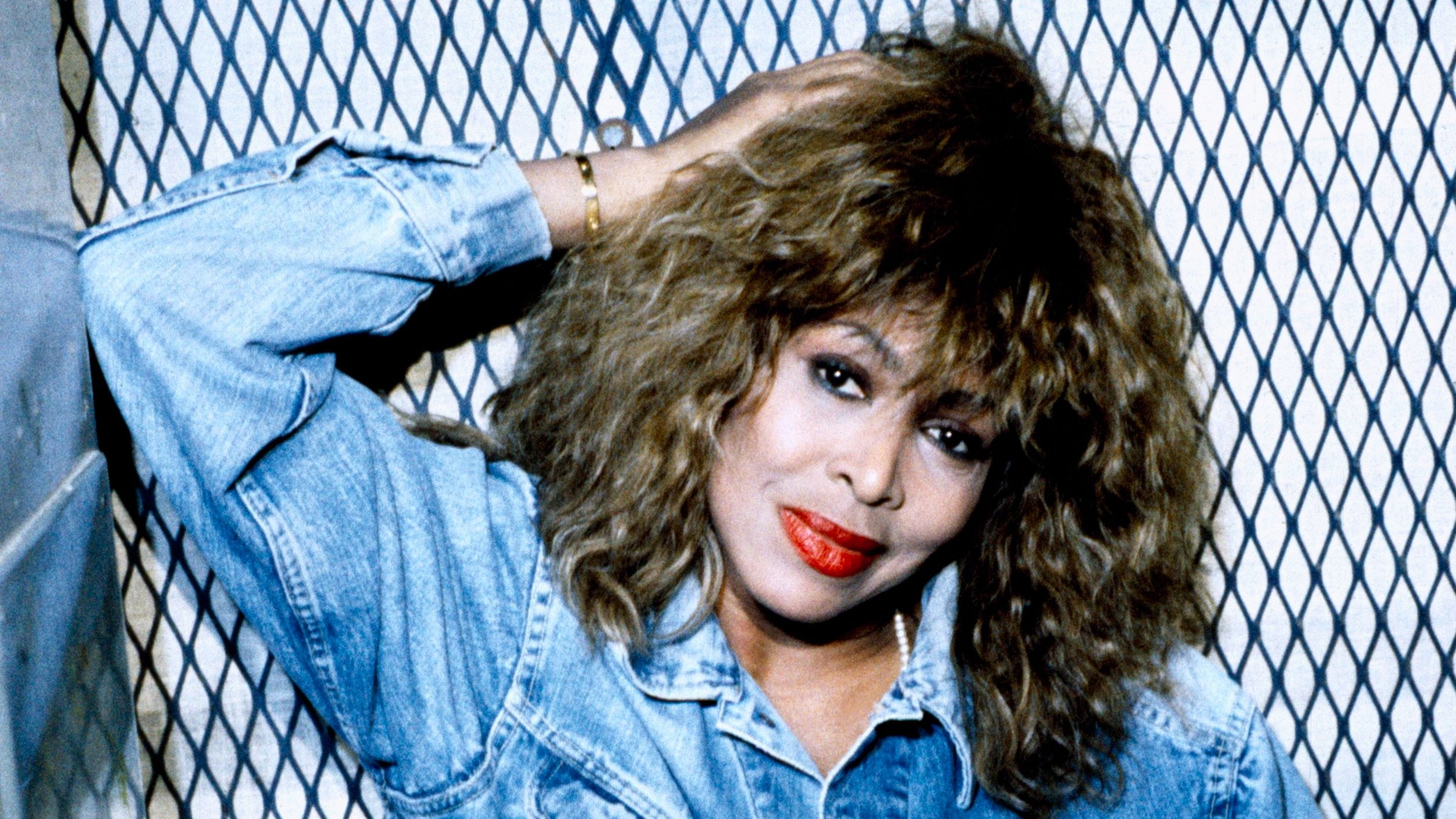 "Tina Turner: Foreign Affair - Live from Barcelona": Tina Turner stützt sich mit dem Ellenbogen seitlich an der Wand ab und hält mit der Hand ihren Kopf. Sie trägt eine blaue Jeansjacke und schaut direkt in die Kamera.