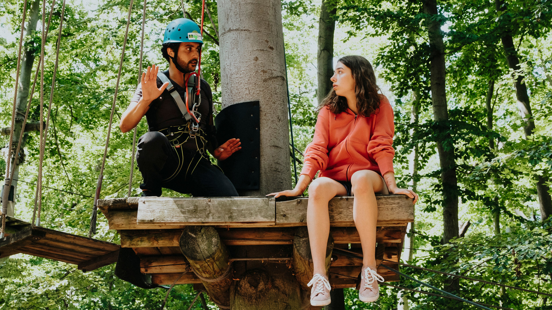 "SOKO Potsdam - Mutterliebe": Fine Nowak (Emilia Eidt) sitzt auf einer hohen Plattform aus Holz im Kletterwald. Sie sieht nach links zu David Grünbaum (Omar El-Saeidi), der neben ihr auf der Plattform hockt. Er ist mit einem Seil an einem Karabinerhaken gesichert, trägt einen blauen Helm und hat beschwichtigend seine Hände gehoben. Fine Nowak ist komplett ungesichert.