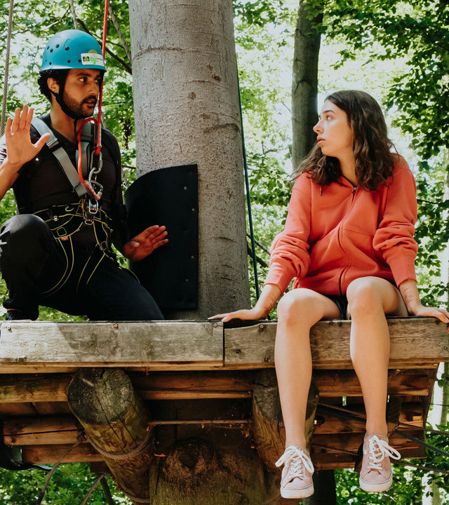 "SOKO Potsdam - Mutterliebe": Fine Nowak (Emilia Eidt) sitzt auf einer hohen Plattform aus Holz im Kletterwald. Sie sieht nach links zu David Grünbaum (Omar El-Saeidi), der neben ihr auf der Plattform hockt. Er ist mit einem Seil an einem Karabinerhaken gesichert, trägt einen blauen Helm und hat beschwichtigend seine Hände gehoben. Fine Nowak ist komplett ungesichert.
