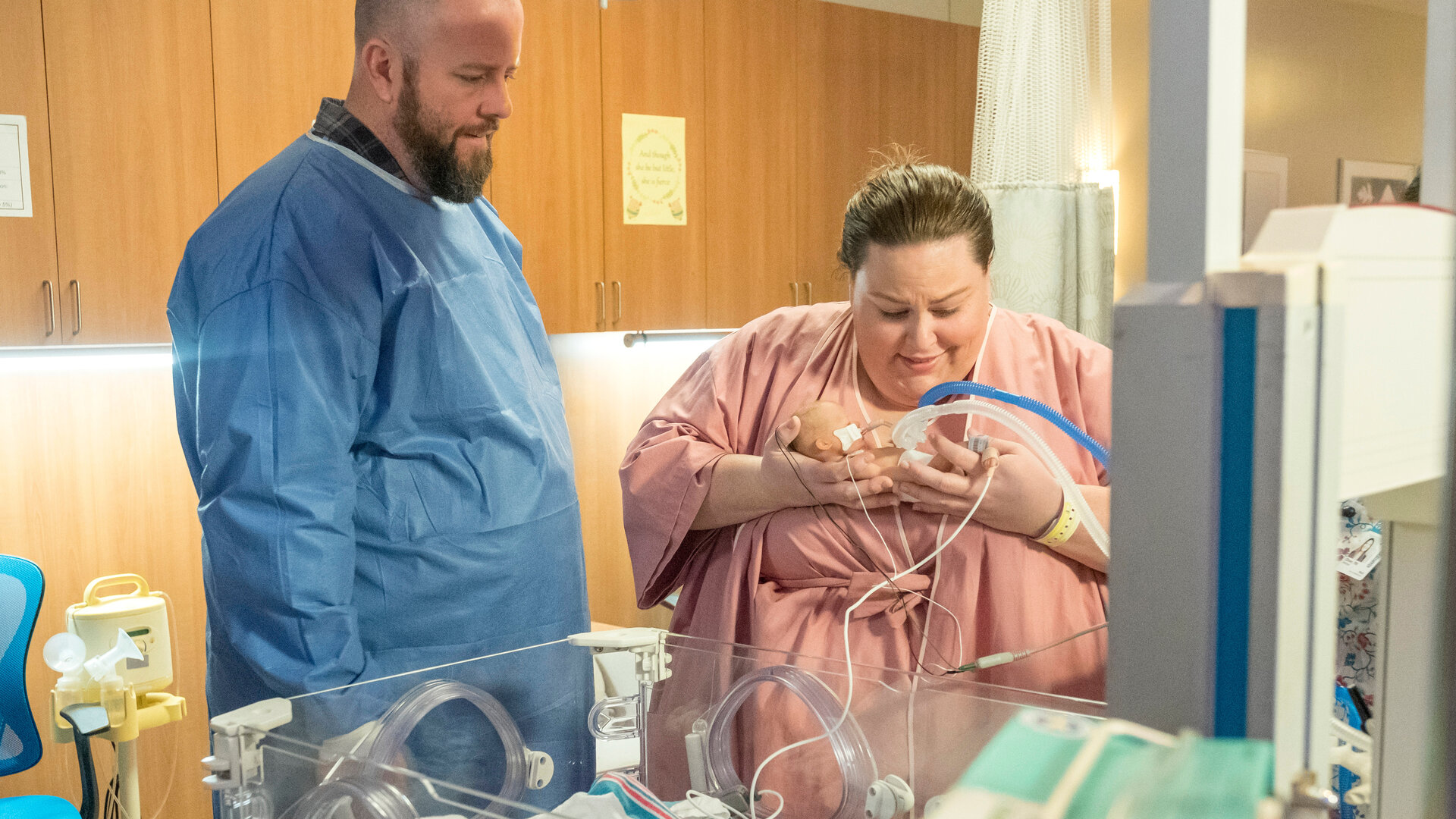 "This Is Us - Das ist Leben: Beziehungsfragen": Kate (Chrissy Metz) und Toby (Chris Sullivan) stehen nebeneinander auf der Neugeborenenstation vor einem leeren Inkubator. Kate trägt ein rosafarbenes Krankenhaushemd und hält ihr Neugeborenes behutsam in den Armen. Das Baby ist an Schläuche und Kabel angeschlossen, die zur medizinischen Überwachung dienen. Toby steht dicht neben ihr und trägt ein blaues Schutzhemd. Beide richten ihren Blick auf das Neugeborene und wirken erleichtert, während sie diesen besonderen Moment gemeinsam erleben.
