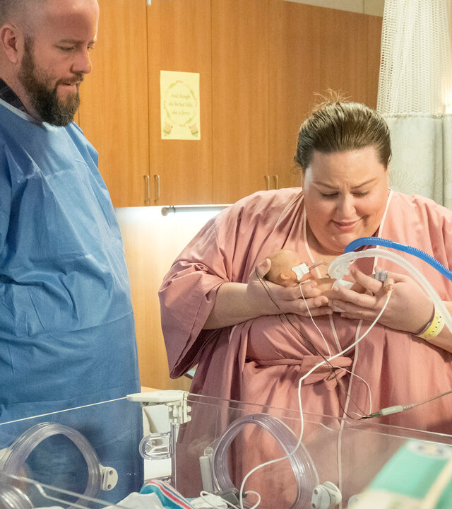 "This Is Us - Das ist Leben: Beziehungsfragen": Kate (Chrissy Metz) und Toby (Chris Sullivan) stehen nebeneinander auf der Neugeborenenstation vor einem leeren Inkubator. Kate trägt ein rosafarbenes Krankenhaushemd und hält ihr Neugeborenes behutsam in den Armen. Das Baby ist an Schläuche und Kabel angeschlossen, die zur medizinischen Überwachung dienen. Toby steht dicht neben ihr und trägt ein blaues Schutzhemd. Beide richten ihren Blick auf das Neugeborene und wirken erleichtert, während sie diesen besonderen Moment gemeinsam erleben.