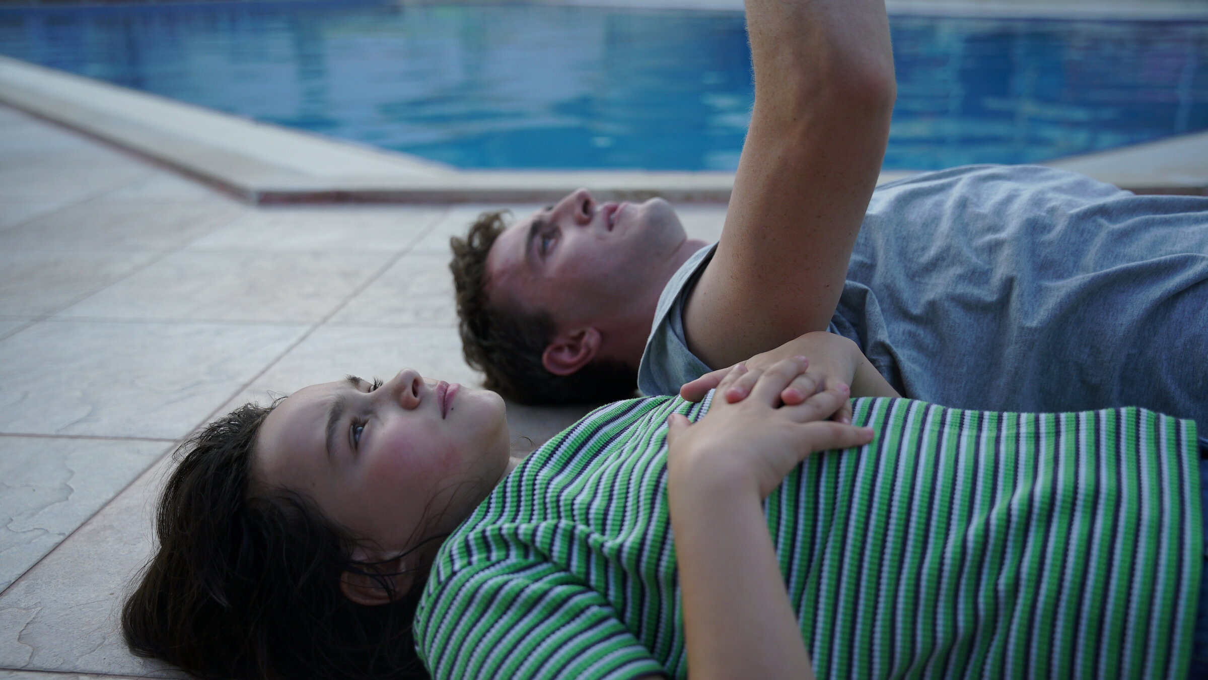 "Aftersun": Sophie (Frankie Corio) und ihr Vater Callum (Paul Mescal) liegen bekleidet mit dem Rücken auf einem gepflasterten Boden, im Hintergrund ist die blaue Wasseroberfläche eines Pools zu sehen.