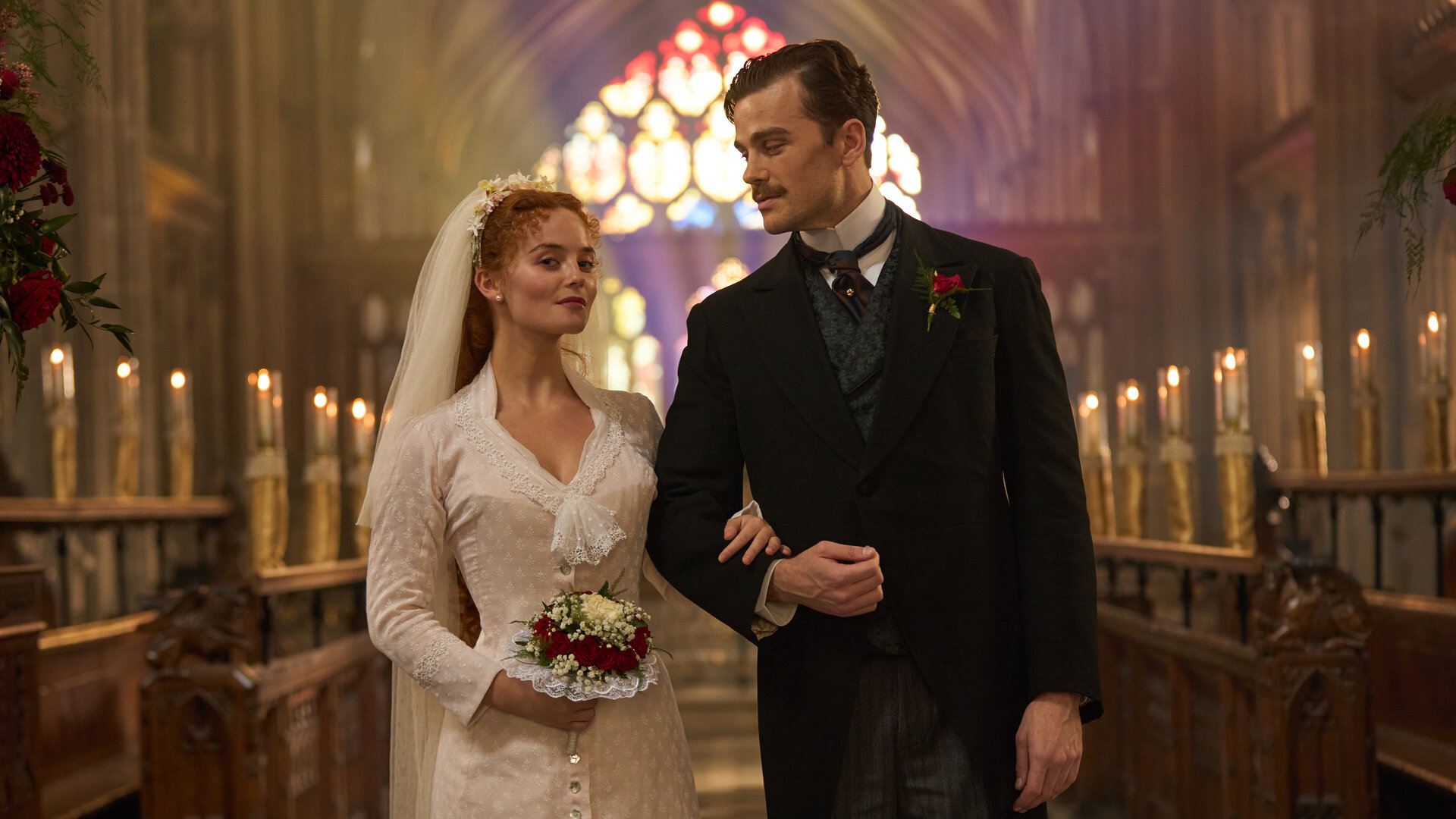 "The Forsytes - Familie verpflichtet: Loyalität oder Liebe": Irene (Millie Gibson) und Soames (Joshua Orpin) stehen nebeneinander in einer Kirche. Im Hintergrund leuchten in einer Reihe aufgestellte Kerzen und, farbiges Sonnlicht fällt durch die bunten Kirchenfenster. Sie trägt ein schlichtes weißes Kleid mit einem weißen langen Schleier im Haar und hält einem Blumenstrauß in der Hand. Soames trägt einen schwarzen Anzug mit Nadelstreifenhose und dazu passender Weste. Irene hat sich bei Soames eingehackt und blickt nach vorne, während er sie ansieht.