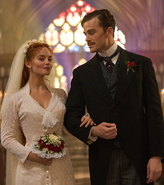 "The Forsytes - Familie verpflichtet: Loyalität oder Liebe": Irene (Millie Gibson) und Soames (Joshua Orpin) stehen nebeneinander in einer Kirche. Im Hintergrund leuchten in einer Reihe aufgestellte Kerzen und, farbiges Sonnlicht fällt durch die bunten Kirchenfenster. Sie trägt ein schlichtes weißes Kleid mit einem weißen langen Schleier im Haar und hält einem Blumenstrauß in der Hand. Soames trägt einen schwarzen Anzug mit Nadelstreifenhose und dazu passender Weste. Irene hat sich bei Soames eingehackt und blickt nach vorne, während er sie ansieht.