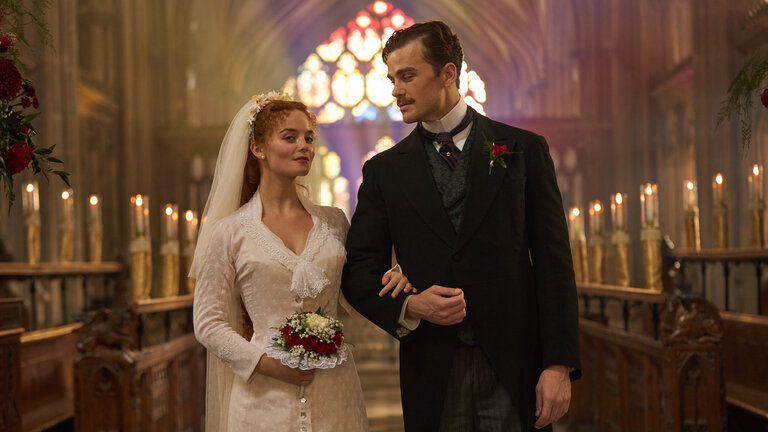 "The Forsytes - Familie verpflichtet: Loyalität oder Liebe": Irene (Millie Gibson) und Soames (Joshua Orpin) stehen nebeneinander in einer Kirche. Im Hintergrund leuchten in einer Reihe aufgestellte Kerzen und, farbiges Sonnlicht fällt durch die bunten Kirchenfenster. Sie trägt ein schlichtes weißes Kleid mit einem weißen langen Schleier im Haar und hält einem Blumenstrauß in der Hand. Soames trägt einen schwarzen Anzug mit Nadelstreifenhose und dazu passender Weste. Irene hat sich bei Soames eingehackt und blickt nach vorne, während er sie ansieht.