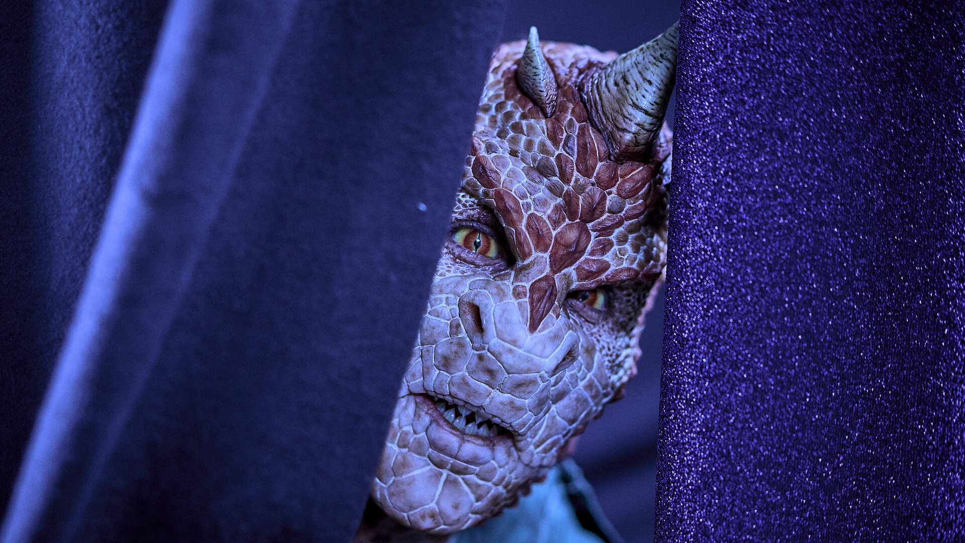 "Club der Dinosaurier - Der Abiball": Hinter einem violetten Vorhang lugt der Kopf eines Teenagers hervor, der sich in einen Dinosaurier verwandelt hat. Er hat große rote Schuppen, rot-gelbe Augen und mehrere Hörner. Die Zähne sind spitz.