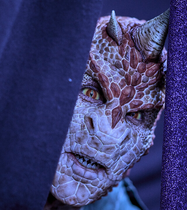 "Club der Dinosaurier - Der Abiball": Hinter einem violetten Vorhang lugt der Kopf eines Teenagers hervor, der sich in einen Dinosaurier verwandelt hat. Er hat große rote Schuppen, rot-gelbe Augen und mehrere Hörner. Die Zähne sind spitz.