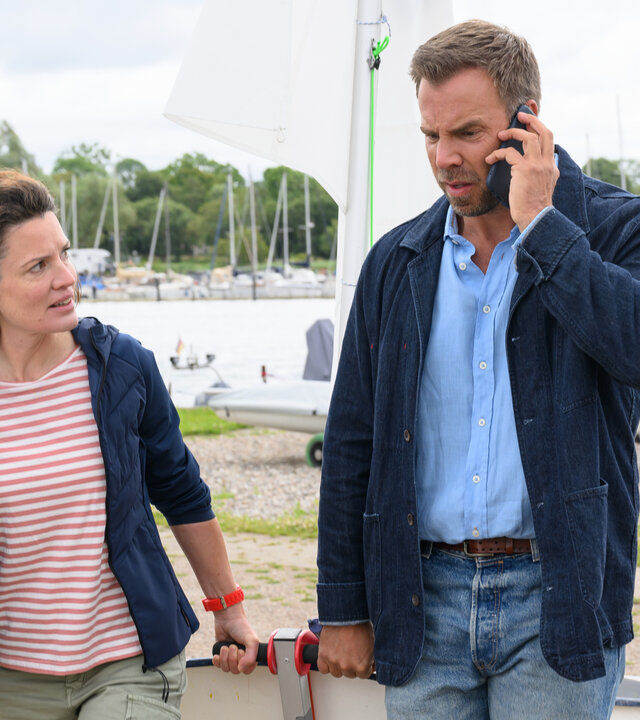 "SOKO Wismar – Scheidewege": Kriminalhauptkommissar Lars Pöhlmann (Dominic Boeer) befragt in einem Segelclub die Schwester des Toten - Segellehrerin Anna Frenzen (Judith Sehrbock). Sie laufen im Freien nebeneinander und ziehen gemeinsam ein Boot. Lars Pöhlmann telefoniert mit dem Handy, Anna Frenzen schaut zu ihm.
