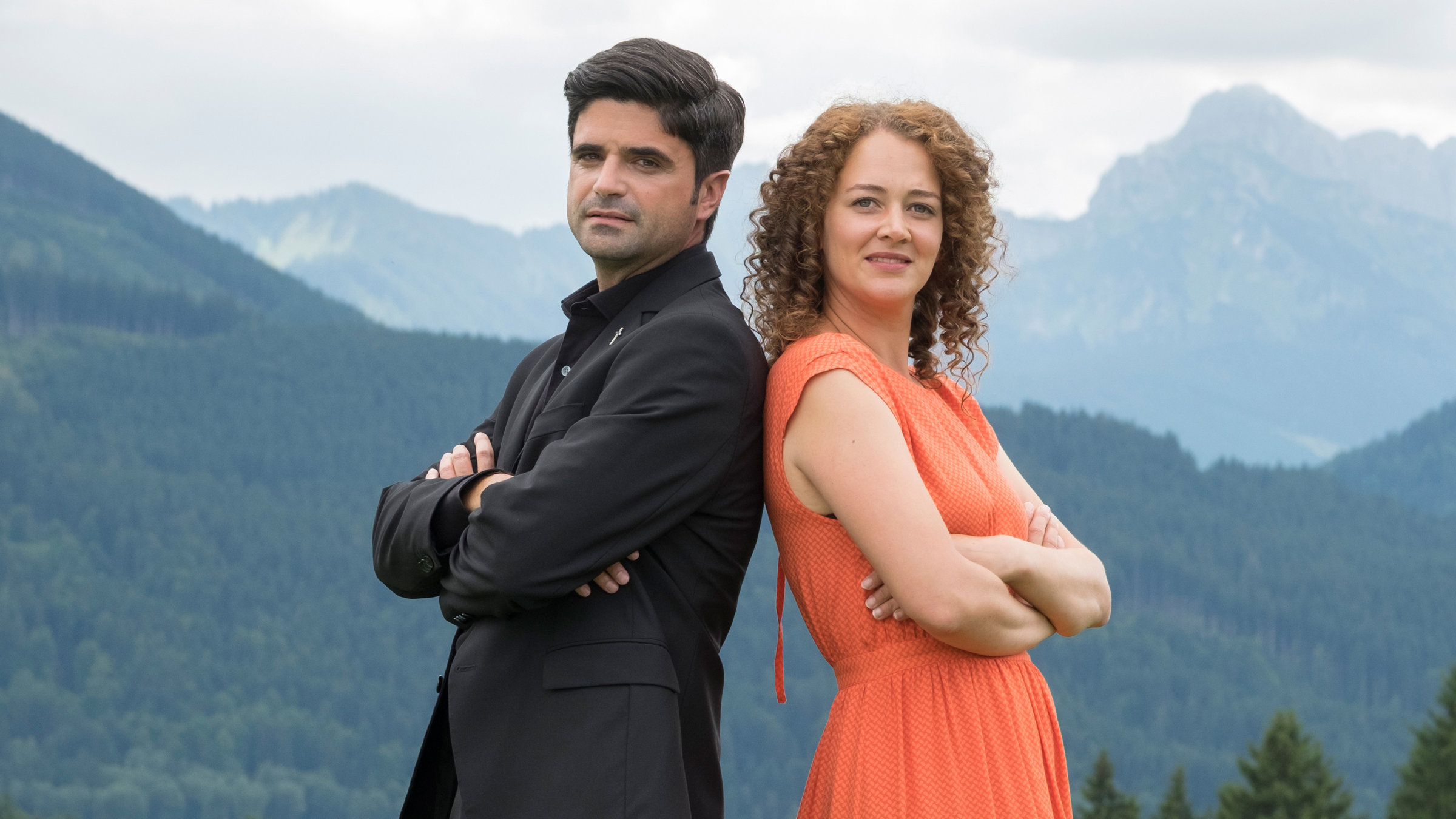 "Tonio & Julia - Zwei sind noch kein Paar": Mit dem Rücken aneinander gelehnt und nach vorne in die Kamera blickend, stehen Pfarrer Tonio Niederegger (Maximilian Grill) und Familientherapeutin Julia Schindel (Oona Devi Liebich) auf einer Wiese. Im Bildhintergrund Bäume und Berge.