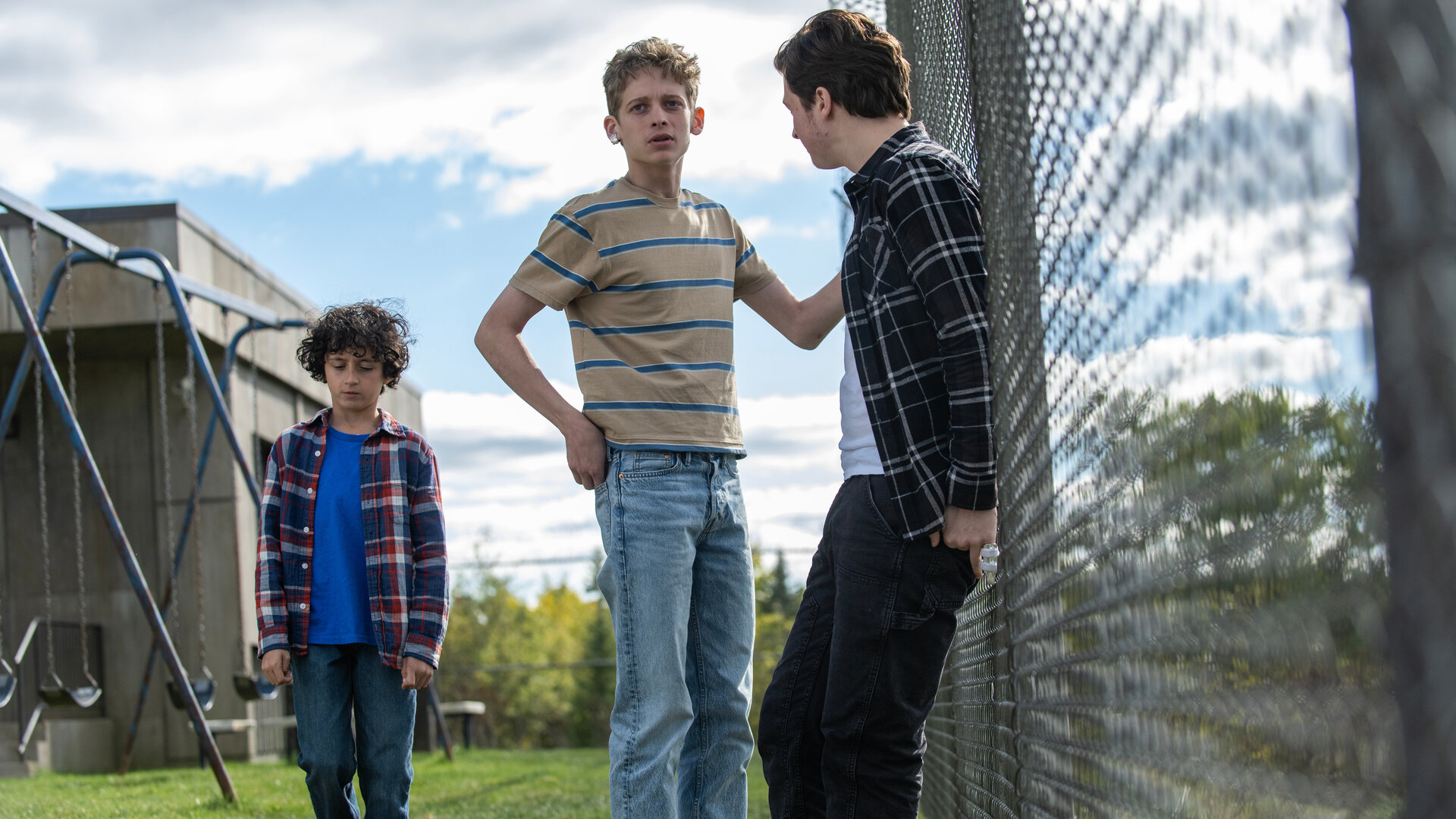 "Das Institut - Das ‘Schlüsselloch‘": Auf dem Spielplatz des Außenbereichs lehnt Nicky (Fionn Laird) mit dem Rücken an einem hohen Maschendrahtzaun und schaut Luke (Joe Freeman) an. Der hält sich mit der linken Hand am Zaun fest und sieht nach vorne. Neben ihm steht der kleine Avery (Viggo Hanvelt). Er blickt traurig nach unten.
