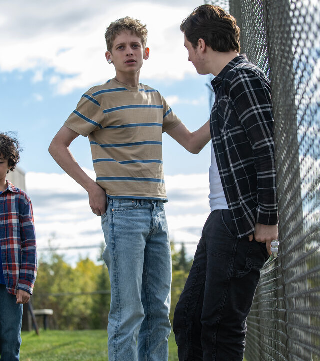 "Das Institut - Das ‘Schlüsselloch‘": Auf dem Spielplatz des Außenbereichs lehnt Nicky (Fionn Laird) mit dem Rücken an einem hohen Maschendrahtzaun und schaut Luke (Joe Freeman) an. Der hält sich mit der linken Hand am Zaun fest und sieht nach vorne. Neben ihm steht der kleine Avery (Viggo Hanvelt). Er blickt traurig nach unten.